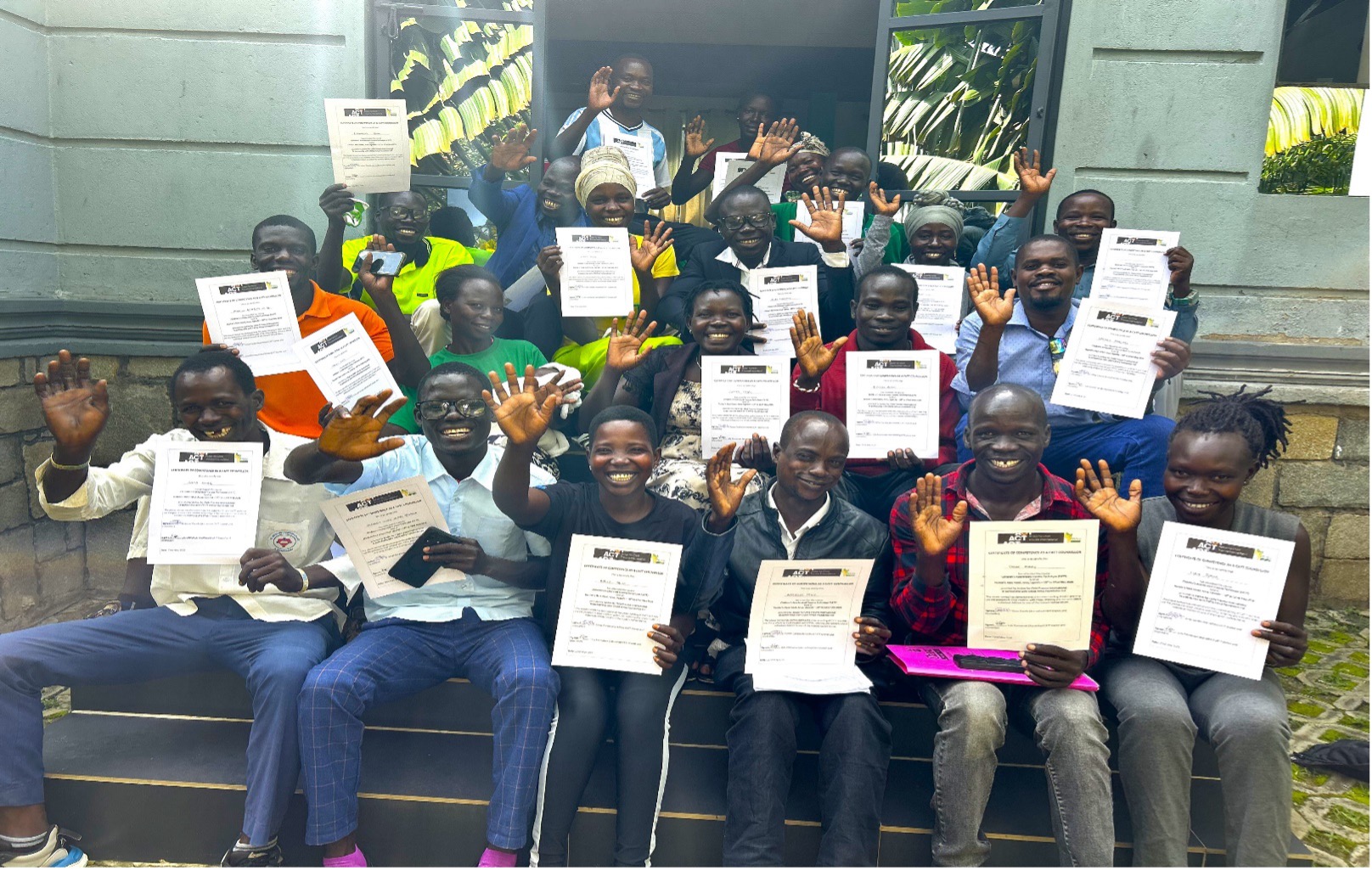 a group of 20 sitting on steps outside green building. They are holding certificates and smiling