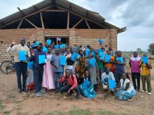 group of men and women standing outside large wooden building. they are holding blue workbooks and laughing/smiling
