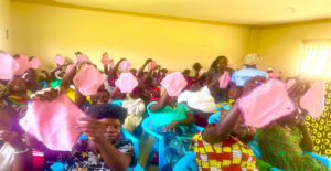 women seated in rows in a yellow room. they are holding up pink handmade menstrual pads. There are 60 women in the room with two trainers in the middle inspecting the pads