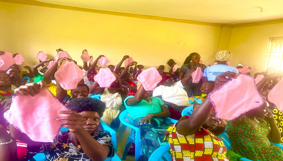 women seated in rows in a yellow room. they are holding up pink handmade menstrual pads. There are 60 women in the room with two trainers in the middle inspecting the pads