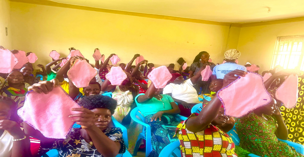 women seated in rows in a yellow room. they are holding up pink handmade menstrual pads. There are 60 women in the room with two trainers in the middle inspecting the pads