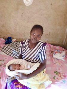 woman sat on a bed with pink floral sheets. she is wearing a purple striped top with purple patterned skirt. She is holding her baby who is swaddled in a pink and yellow cloth