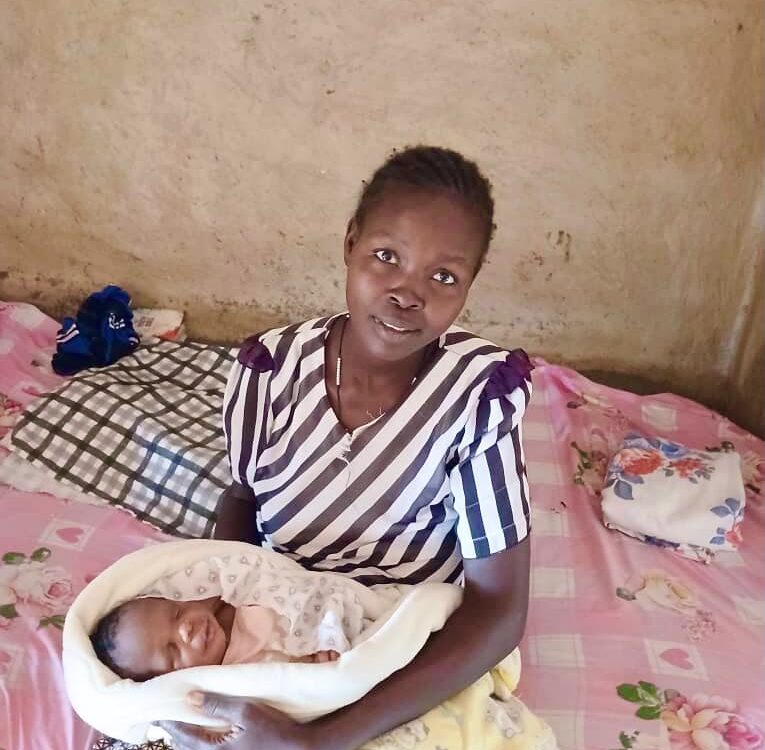 woman sat on a bed with pink floral sheets. she is wearing a purple striped top with purple patterned skirt. She is holding her baby who is swaddled in a pink and yellow cloth