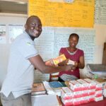 man waring grey polo and trousers is handing yellow box of stationary to a teacher wearing a red blouse. They are in a classroom with learning posters on the wall. There is a pile of red and white stationary boxes on a desk in front of the teacher