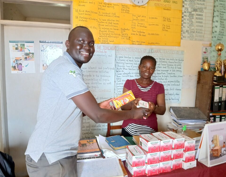 man waring grey polo and trousers is handing yellow box of stationary to a teacher wearing a red blouse. They are in a classroom with learning posters on the wall. There is a pile of red and white stationary boxes on a desk in front of the teacher