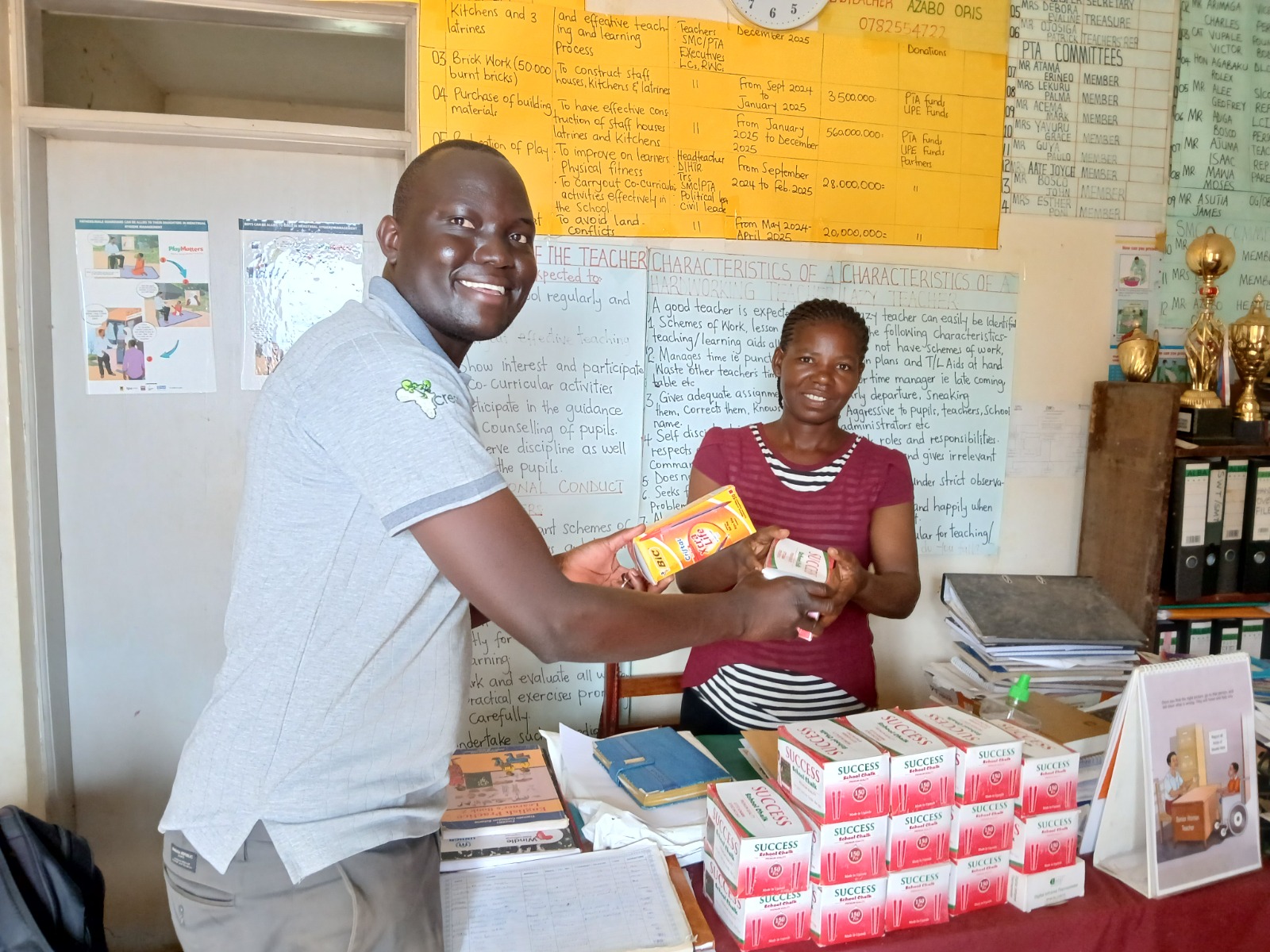 man waring grey polo and trousers is handing yellow box of stationary to a teacher wearing a red blouse. They are in a classroom with learning posters on the wall. There is a pile of red and white stationary boxes on a desk in front of the teacher