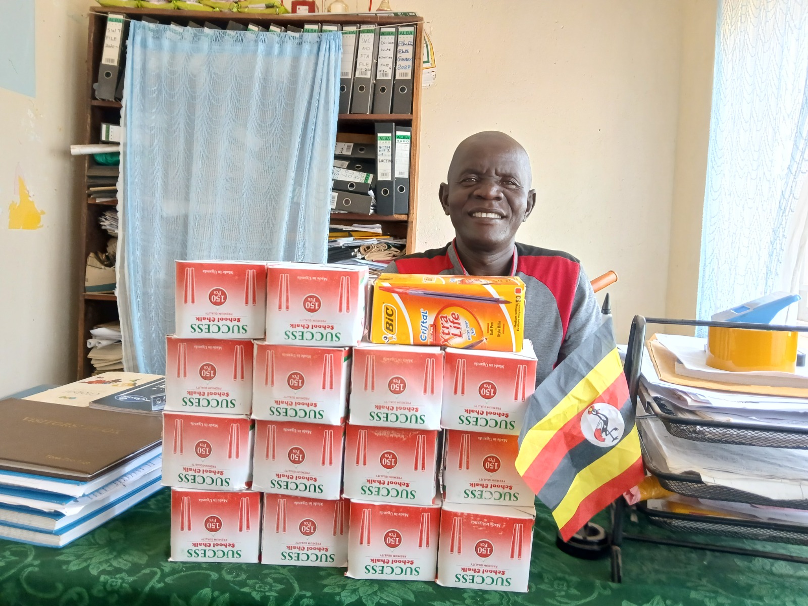 Teacher sits behind a desk. He has a bog smile on his face. Piled up in front of him on the desk are red and white, and yellow boxes of stationary.